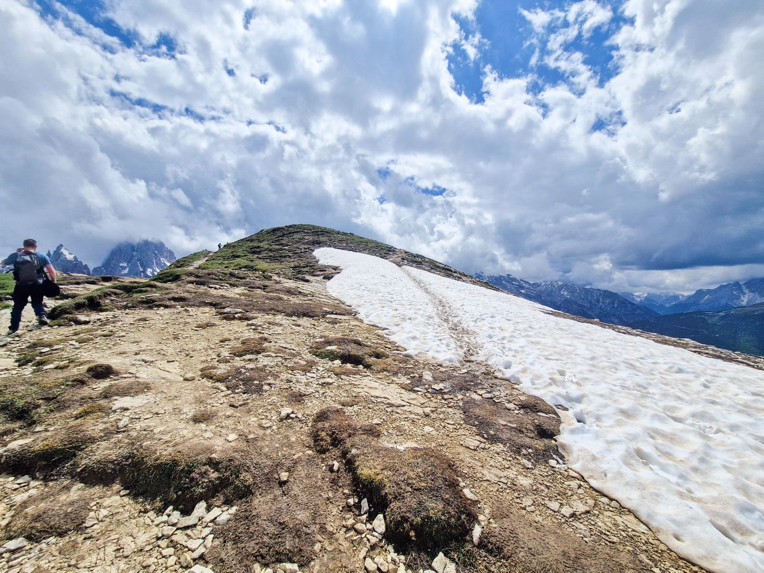 Cadini di Misurina Viewpoint Hike: map + secret spot! 7 A hiker walks up a rocky path toward a snow-covered section of a mountain under a cloudy sky. Snow patches contrast with the bare ground, and distant peaks are visible under the dramatic clouds, capturing the breathtaking essence of the Cadini di Misurina viewpoint hike.