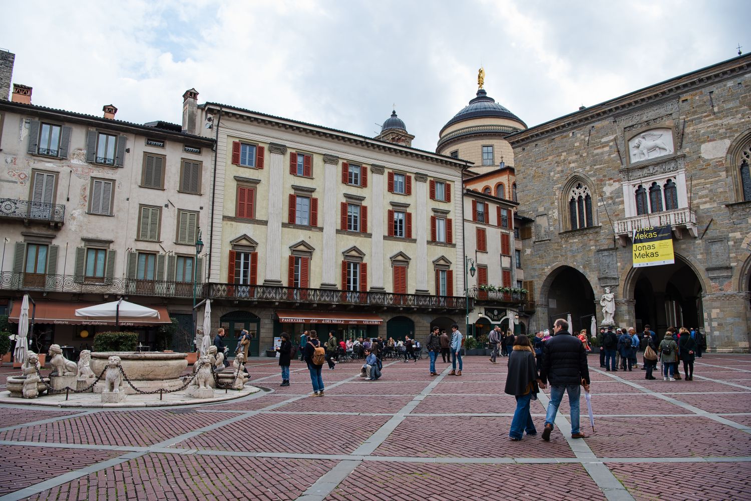 A bustling European square with people walking and gathering. Historic buildings surround the plaza, featuring red and beige facades. A stone fountain is in the foreground, and a weathered church is in the background with a banner visible.