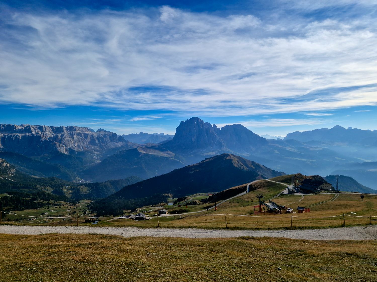11 Short and Easy Hikes in the Dolomites: Unique and Must-do 6 Scenic landscape of rolling hills and valleys with a rugged mountain range in the distance under a cloudy blue sky. The foreground features grassy fields and scattered paths, creating a tranquil, expansive view.
