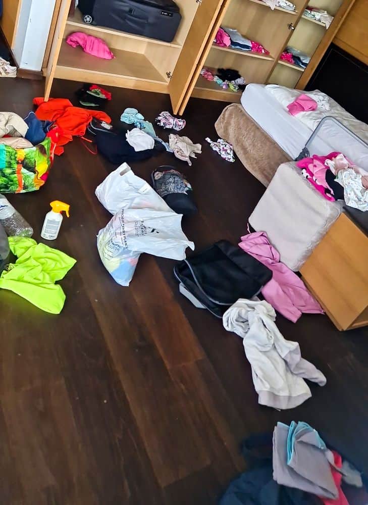 Dolomites Packing List: Plus a Free PDF Printable! 3 A messy room with clothes scattered on a dark wooden floor. An open wardrobe holds more clothes and luggage. A bed with a beige blanket is partially visible, with bags and garments strewn around.