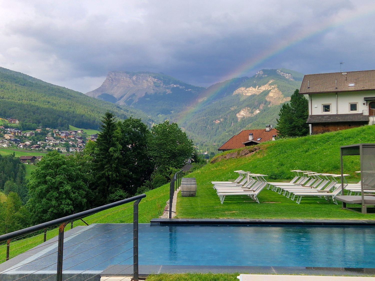 An outdoor infinity pool overlooks a green mountain valley beneath dark storm clouds and a faint rainbow stretching across the sky. White lounge chairs line the grassy hillside while scattered houses sit among the forested hills and rocky peaks in the distance.
