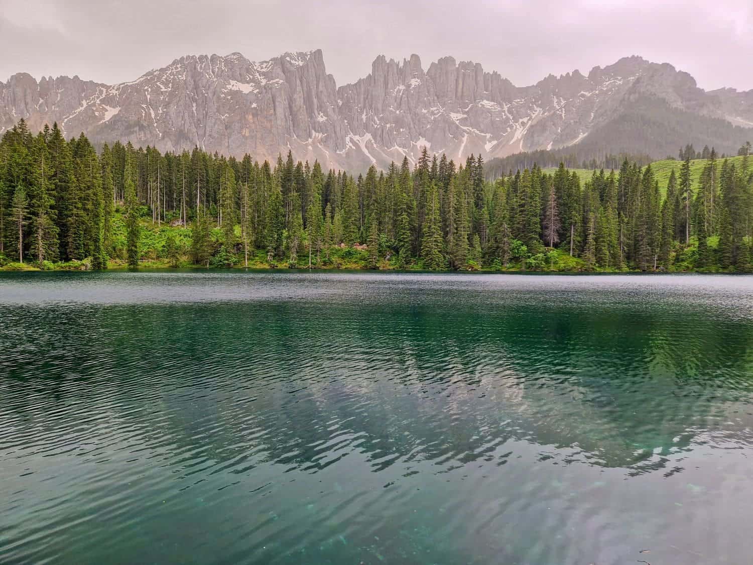 Lago di Carezza: Visit The Rainbow Lake of the Dolomites 7 A serene lake carezza with calm, reflective waters is surrounded by dense, tall pine trees. In the background, jagged gray mountains partially covered with patches of snow rise into a cloudy sky, creating a peaceful and picturesque landscape.