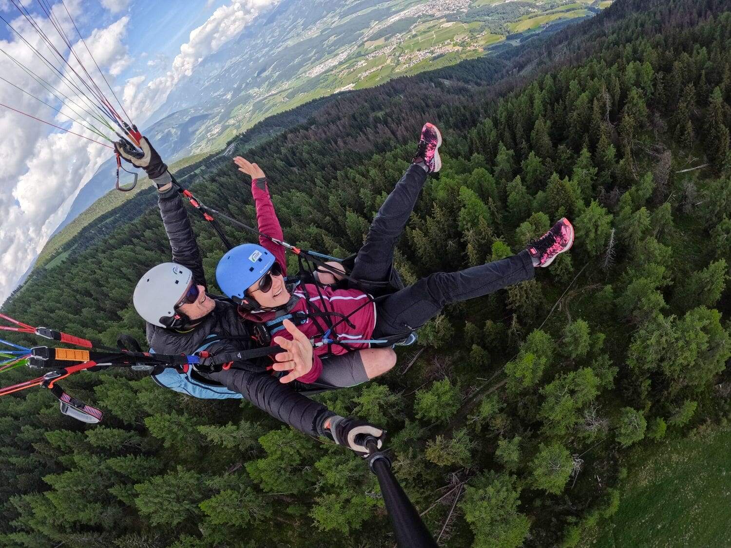 Paragliding in the Dolomites Italy: Complete Guide 5 Two people are paragliding in the Dolomites over a lush forest, with one person wearing a blue helmet and the other a red helmet. They are tandem paragliding, with the instructor in the rear. Both have their arms outstretched, and a scenic landscape of trees and hills is visible below.