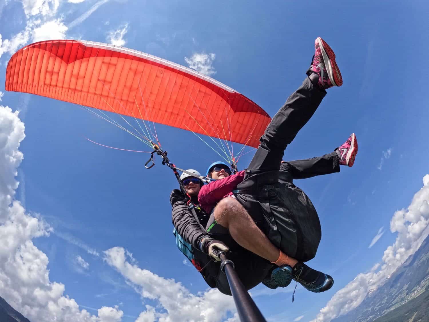 Paragliding in the Dolomites Italy: Complete Guide 4 Two people are tandem paragliding high in the sky with a red paraglider. They are wearing helmets, harnesses, and sunglasses, soaring above a landscape of green hills under a blue sky scattered with white clouds. Paragliding in the Dolomites, they appear to be enjoying the adventure immensely.