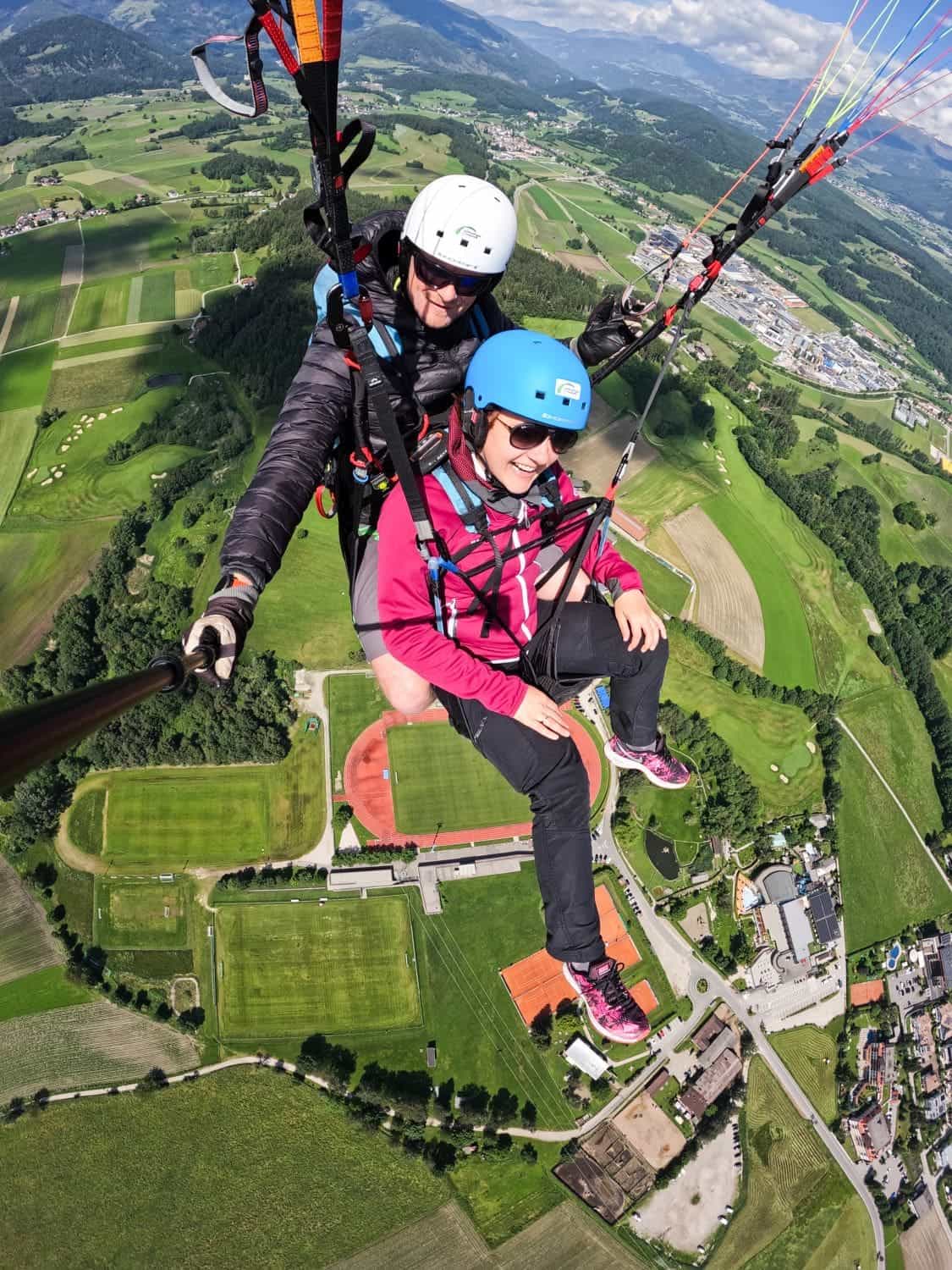 Paragliding in the Dolomites Italy: Complete Guide 3 Two individuals are tandem paragliding high above a picturesque landscape of green fields, roads, and buildings in the Dolomites. Both are wearing helmets and harnesses; the back person is holding a selfie stick to capture the moment. The sky is blue with scattered clouds, and mountains are visible in the distance.