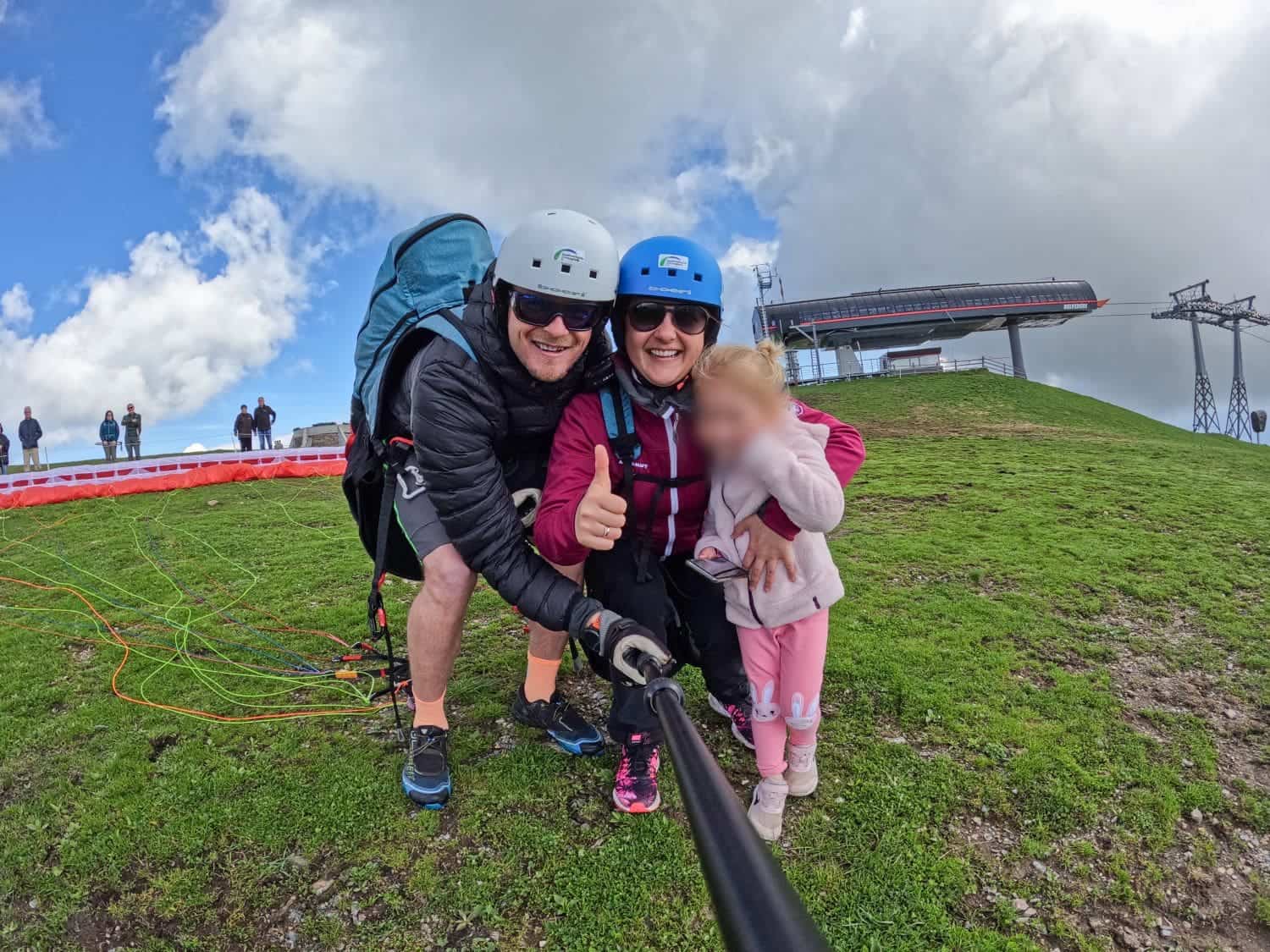 Paragliding in the Dolomites Italy: Complete Guide 2 Three people on a grassy hilltop in the Dolomites, preparing for a paragliding adventure. The adults are wearing helmets and harnesses, smiling at the camera. The child, whose face is blurred, is holding onto one adult. A ski lift structure and a few people are in the background.