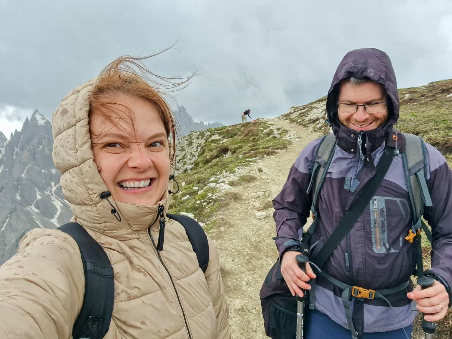 Val Gardena vs Cortina: Where to stay? 14 Two people dressed in outdoor clothing, including jackets and backpacks, smile and pose on a narrow mountain trail. The woman on the left has windblown hair, and the man on the right holds trekking poles. Rugged, cloudy mountain scenery is visible in the background at Cadini di Misurina Viewpoint.