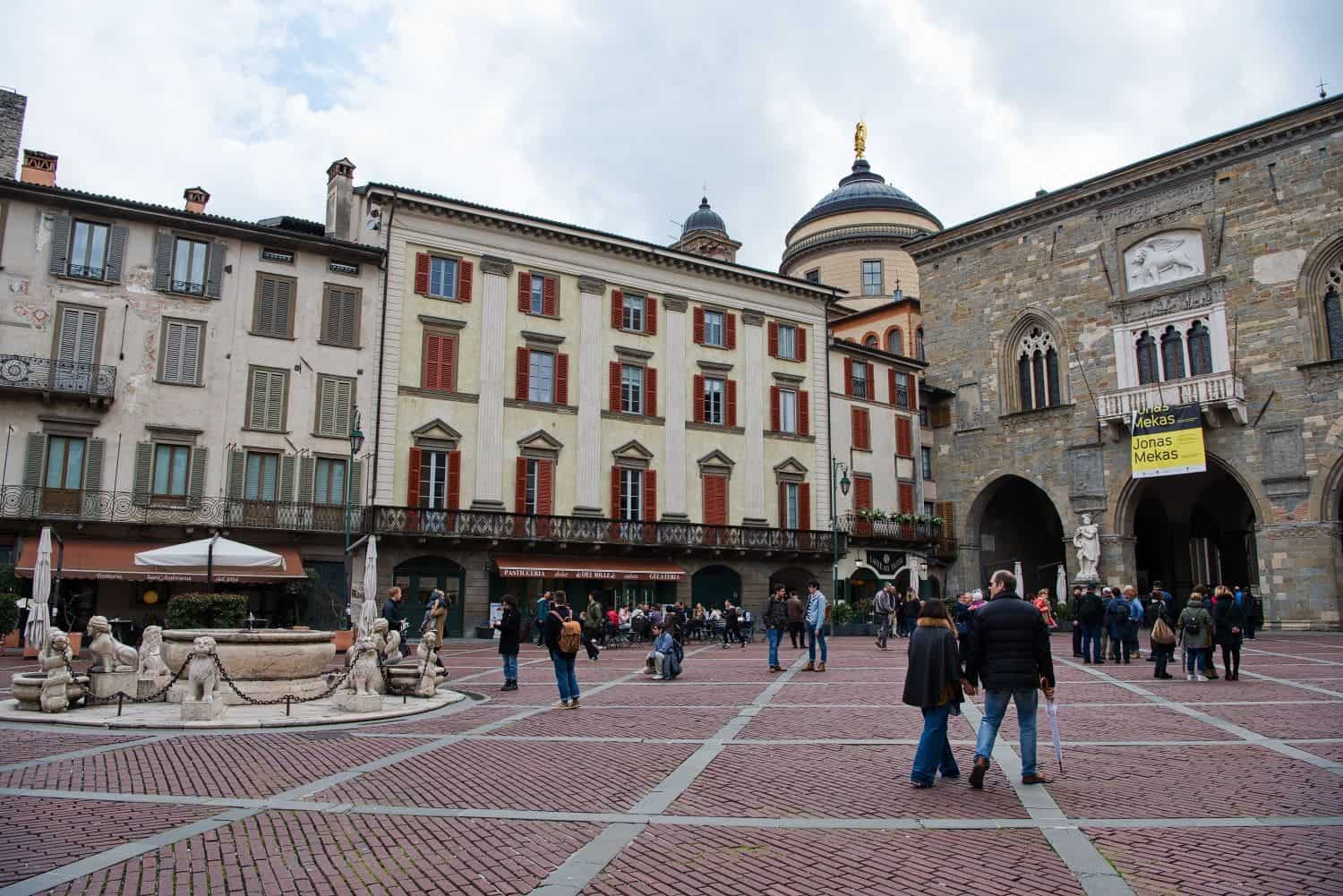 A bustling European square with people walking and gathering. Historic buildings surround the plaza, featuring red and beige facades. A stone fountain is in the foreground, and a weathered church is in the background with a banner visible.