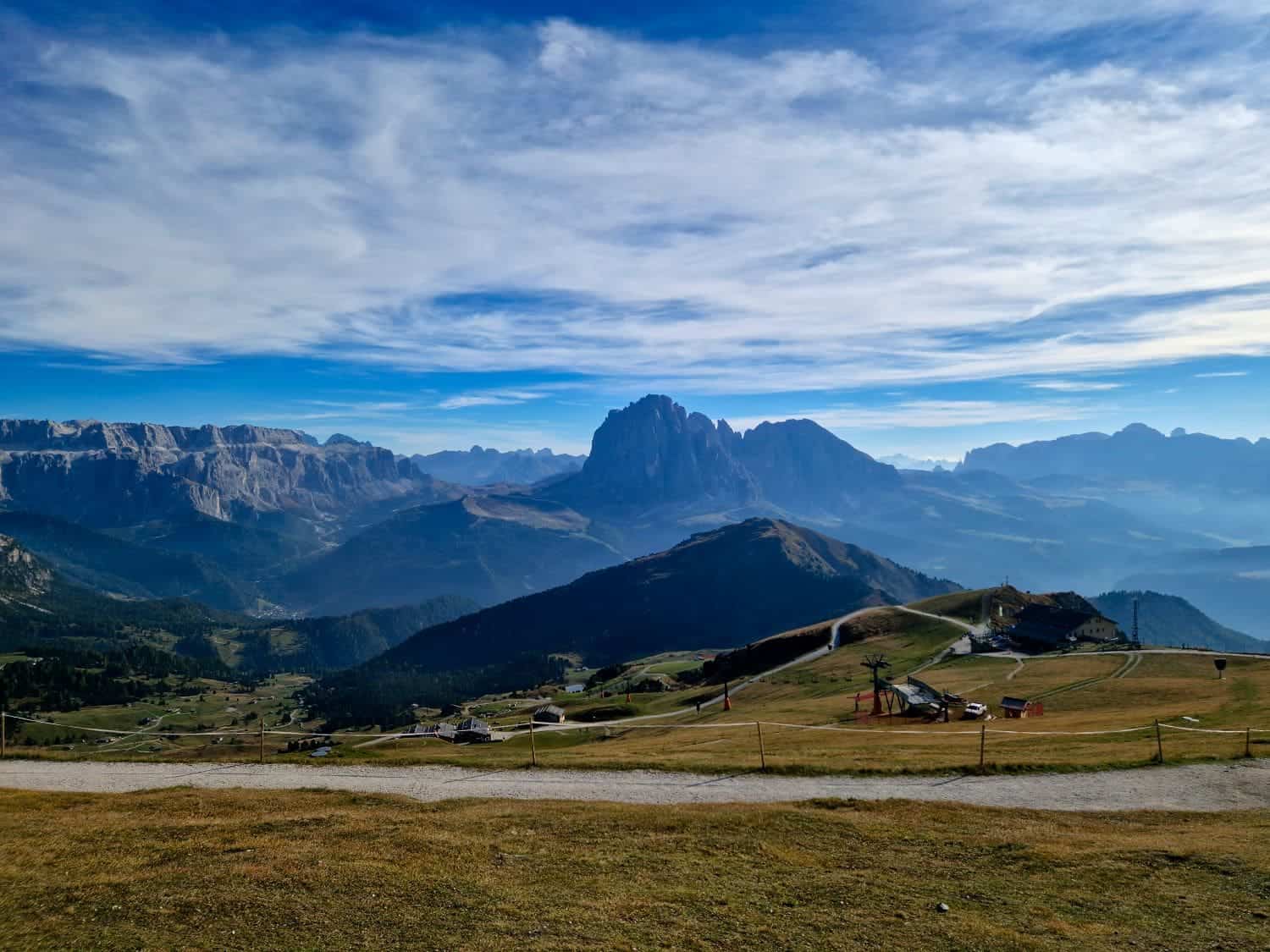 Scenic landscape of rolling hills and valleys with a rugged mountain range in the distance under a cloudy blue sky. The foreground features grassy fields and scattered paths, creating a tranquil, expansive view.