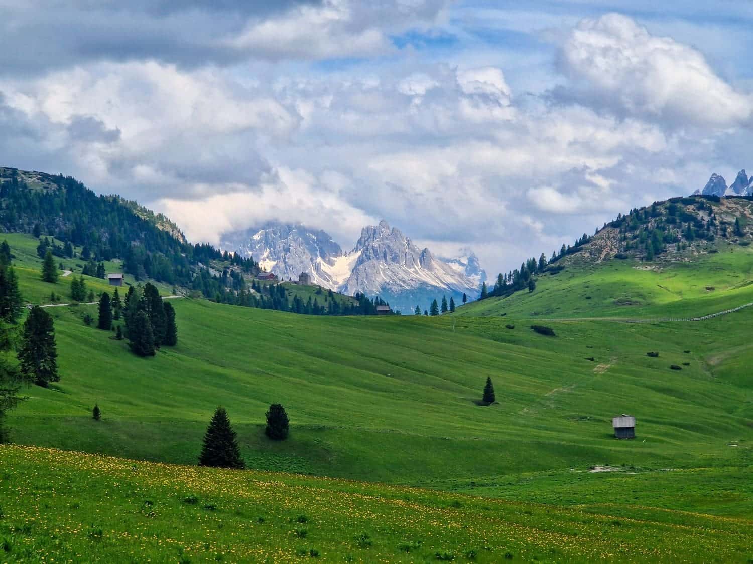 23 Most Beautiful Places in the Dolomites: Including a Map! 24 A scenic landscape featuring a lush green valley with scattered trees and a few huts. Rolling hills lead to distant, snow-capped mountains under a partly cloudy sky, creating a serene and picturesque view.