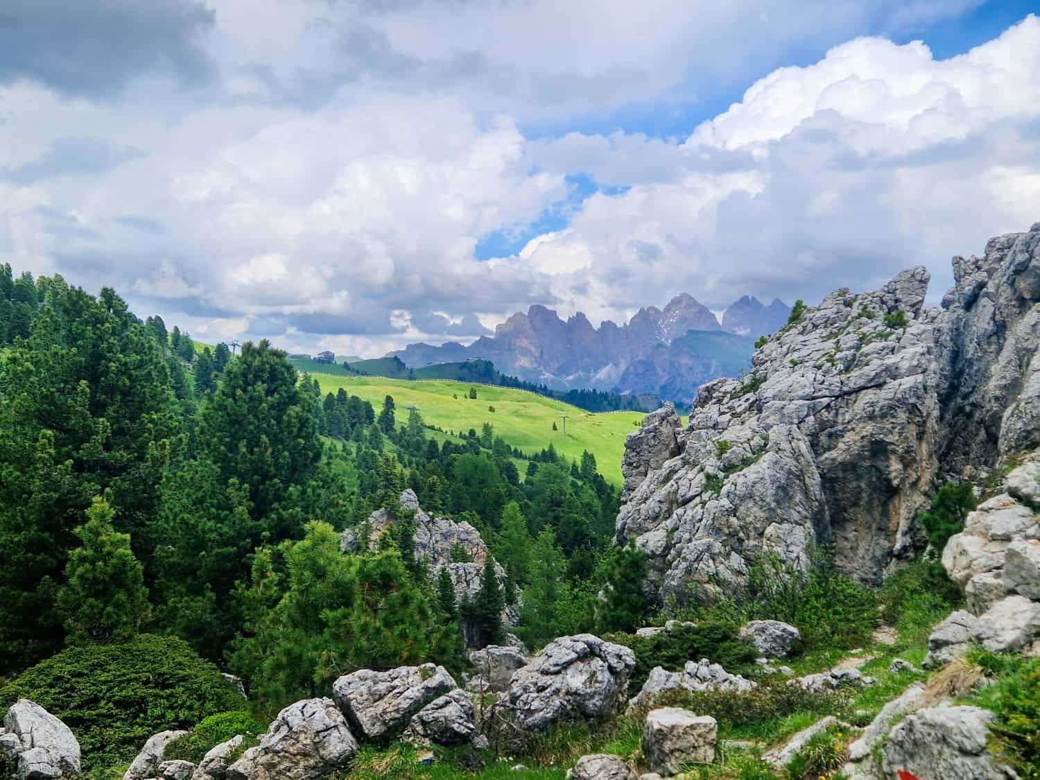Dolomites in June: itinerary and how to guide 69 Scenic view of the Dolomites with a mix of rocky terrain and lush green hills. Cloudy sky above creates dramatic lighting, and distant mountains are partly visible in the background. Large rocks and pine trees frame the landscape.