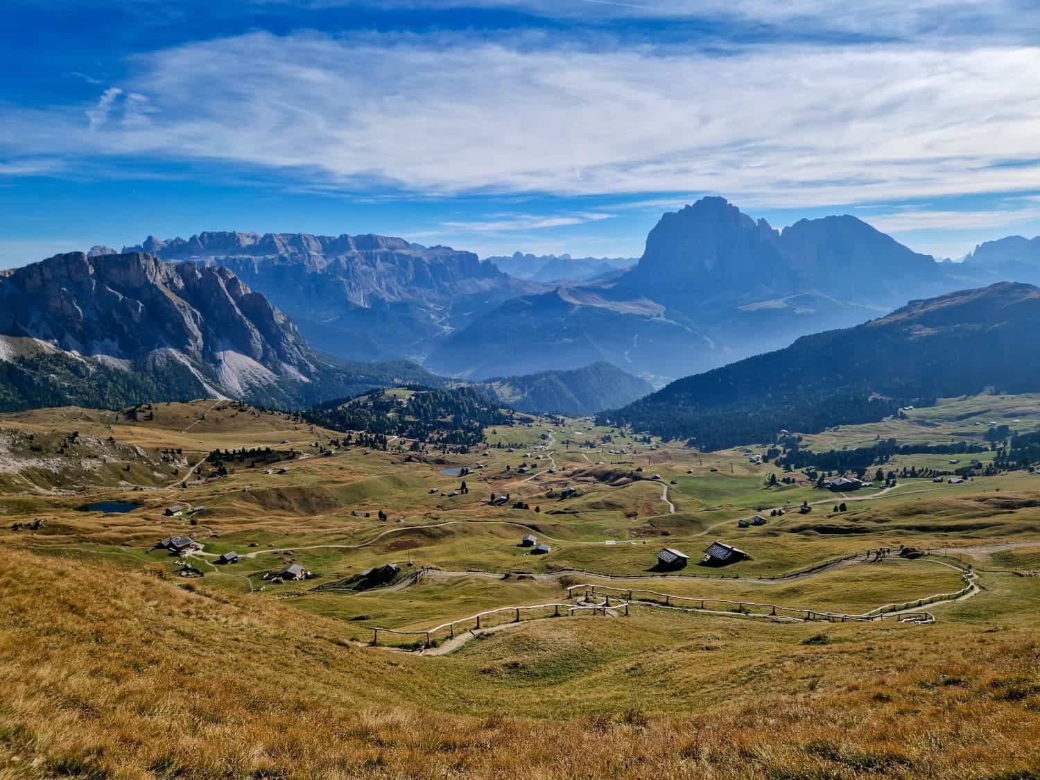 Dolomites in October: is it the best time to go? 8 Scenic view of a vast valley surrounded by mountains under a blue sky with wispy clouds. The landscape is dotted with small cabins and winding paths, with grassy fields and rocky mountain peaks in the distance.