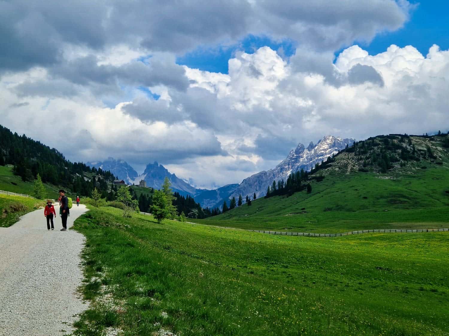 A group of hikers walk along a gravel path through a lush green meadow, surrounded by hills and scattered trees. Snow-capped mountains and a dramatic, cloudy sky form the backdrop.