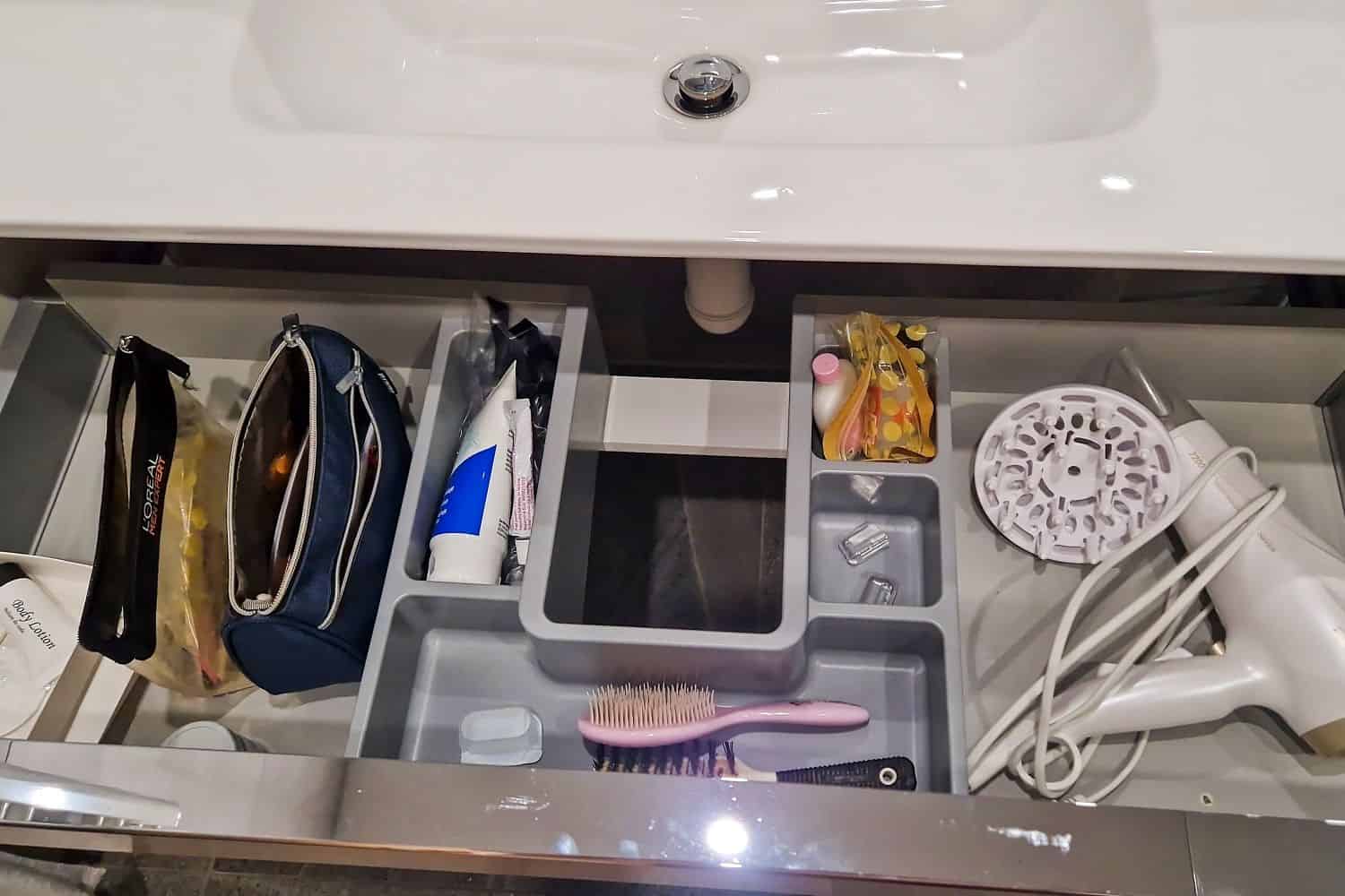 Northern Italy Winter packing list: From Essentials to Specials 12 An open bathroom drawer beneath a sink, organized with various compartments. It contains toiletry bags, a tube of toothpaste, a hairbrush, a hairdryer, and small containers. The drawer is neatly arranged for easy access to personal care items.