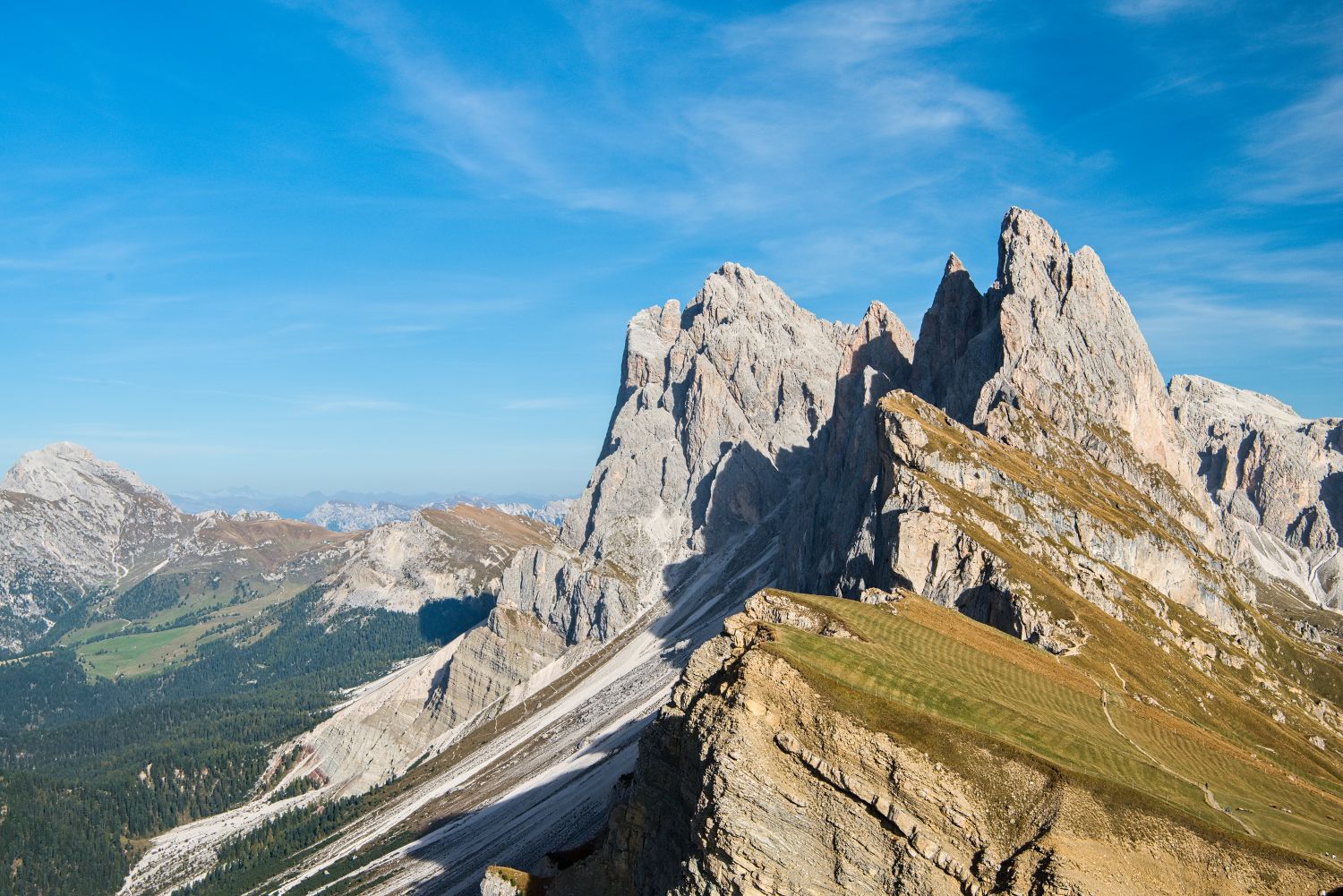 Dolomites in October: is it the best time to go? 7 Mountain landscape with jagged rocky peaks, grassy slopes, and patches of snow under a clear blue sky. Rolling hills and distant mountains are visible in the background.