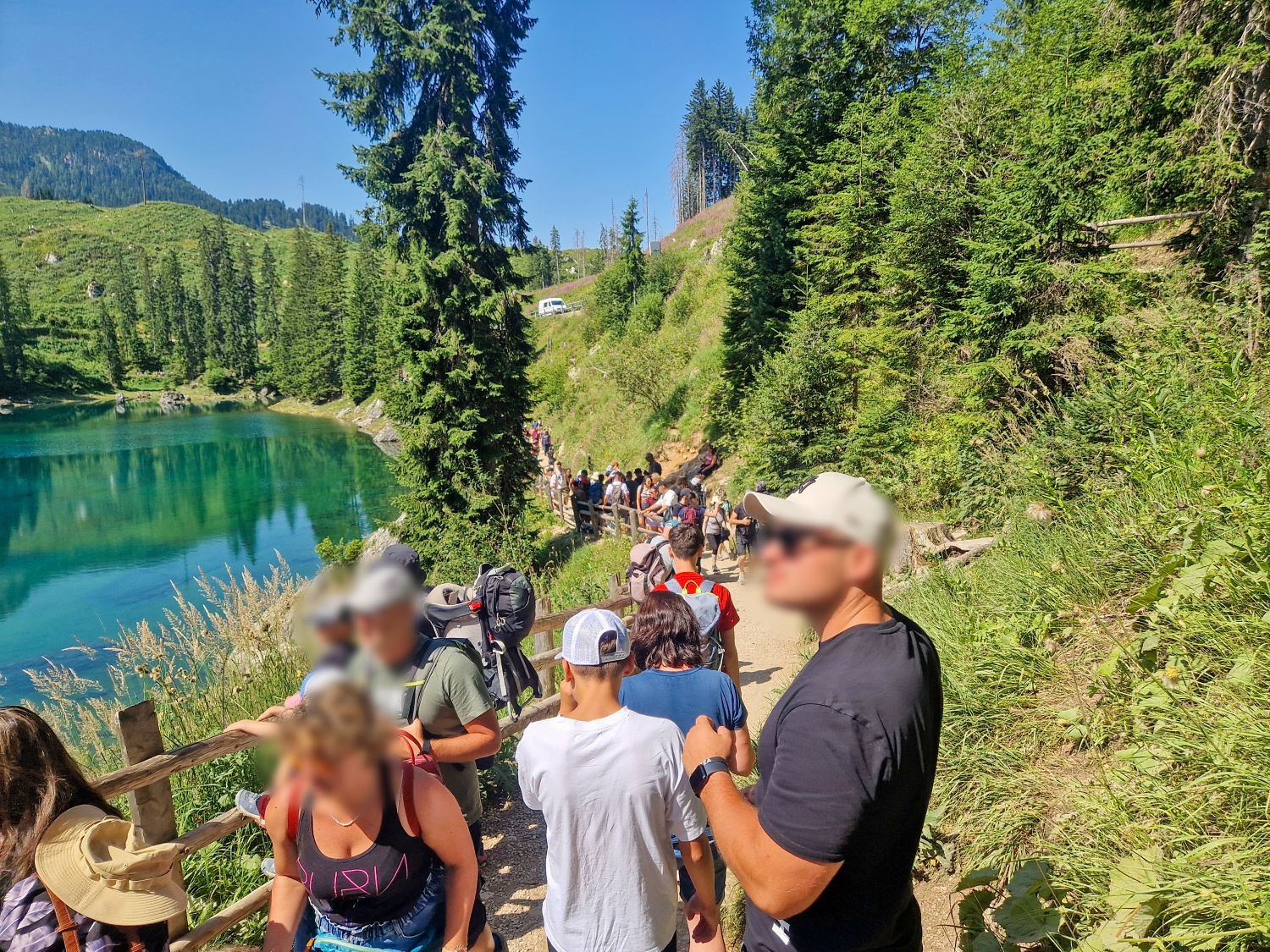 Should you go to the Dolomites in July or August? 8 A group of people, some with backpacks and hats, walk along a narrow dirt trail beside a clear lake surrounded by green hills and tall trees under a bright blue sky.