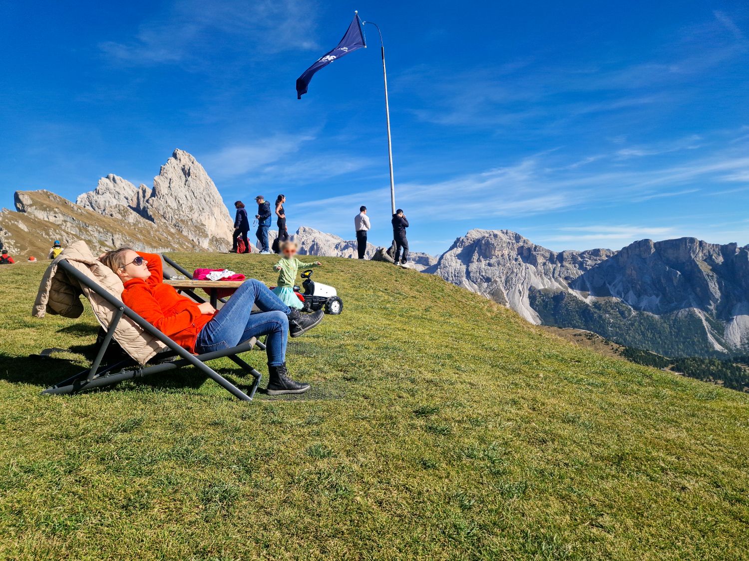 Dolomites in October: is it the best time to go? 19 A person relaxes on a lounge chair on a grassy mountain slope, while others walk nearby. A flag waves on a tall pole. Rocky mountains and a blue sky are in the background.