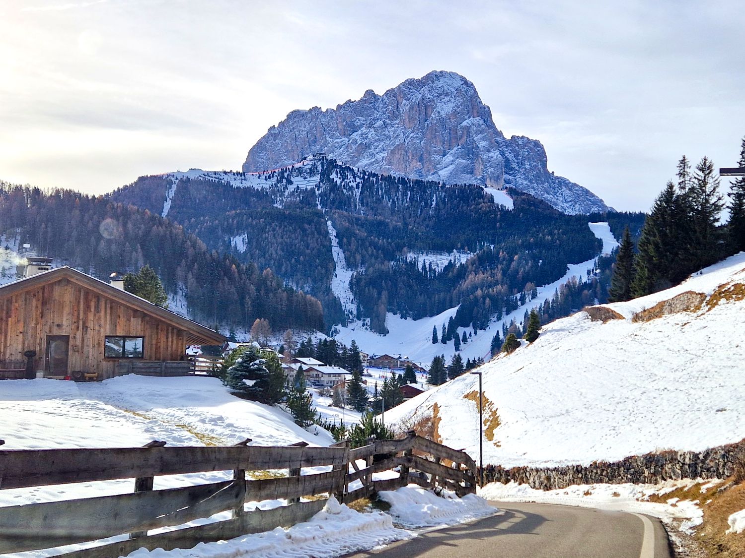 Skiing in Val Gardena and Alpe di Siusi: Ultimate Guide to Italy's Best Ski Resort 2 A quiet mountain road curves through snow covered hills toward a small alpine village, with a wooden chalet in the foreground. A dramatic rocky mountain rises in the background above forested slopes and visible ski runs, creating a peaceful winter landscape.