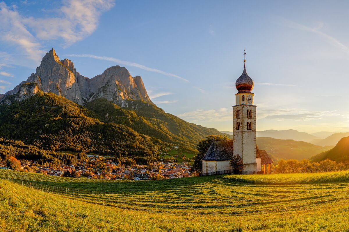 Sunlit summer view of Castelrotto with traditional alpine buildings, green meadows, and rolling hills set against dramatic mountain peaks. The village sits peacefully in the landscape, highlighting the contrast between manicured fields and rugged Dolomite scenery.