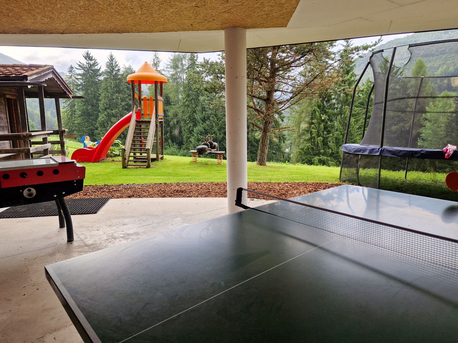 Hotel Digon in Ortisei Review: Best Views or Bad Location? 8 A covered outdoor play area features a ping pong table, foosball table, trampoline, and colorful playground slide surrounded by tall pine trees. The grassy yard overlooks a forested mountain landscape, creating a quiet family-friendly recreation space.